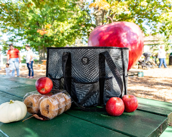 Black mesh bag on a picnic table with apples and donuts, people in the background at an apple orchard.