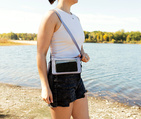 Person standing by a lake wearing a white tank top and black shorts with a crossbody bag.