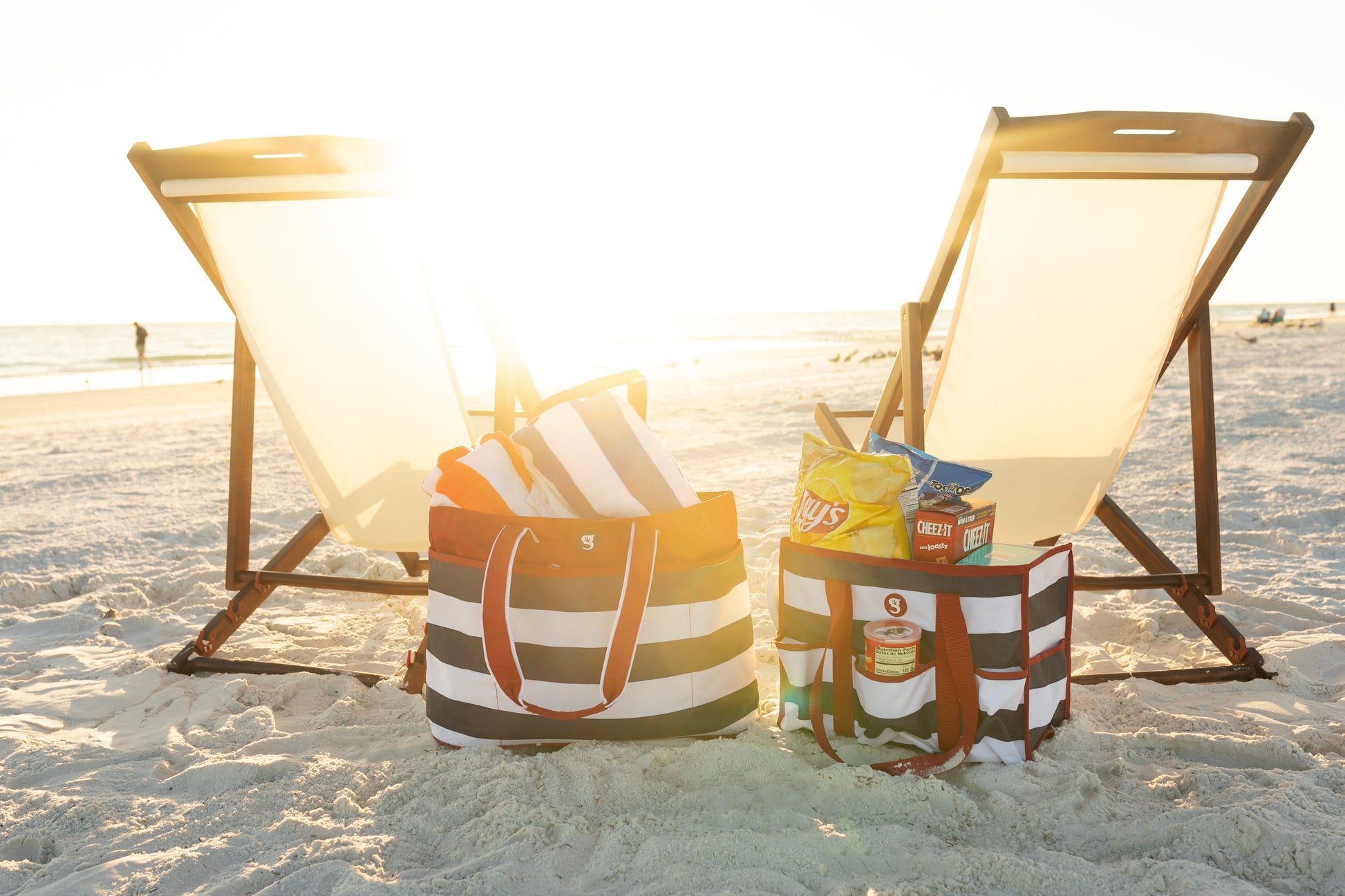 Two beach bags with snacks on a sandy beach with lounge chairs.