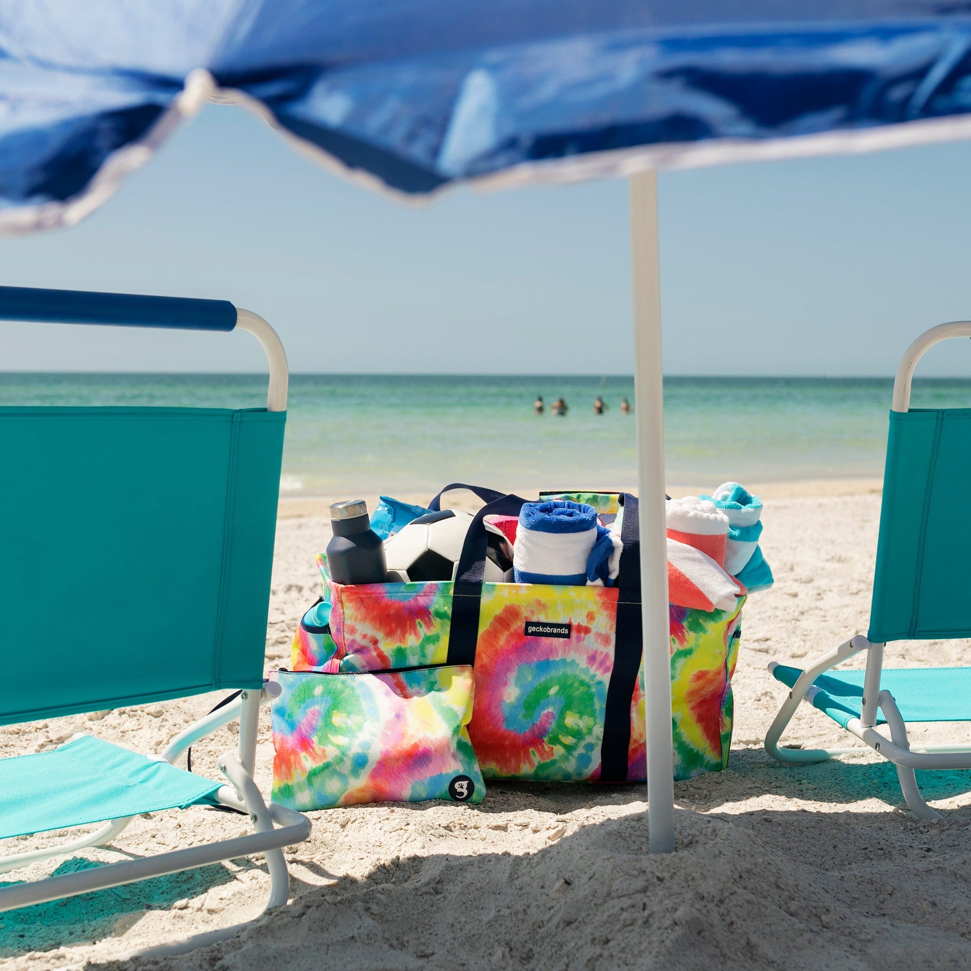 Two turquoise beach chairs under a blue umbrella on a sandy beach with tie-dye bags nearby.