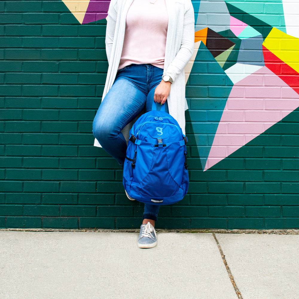 Person holding a blue backpack against a colorful geometric mural on a brick wall.