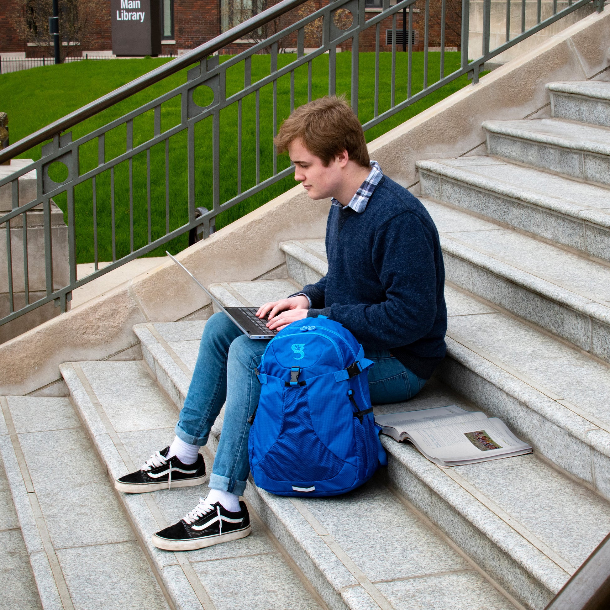 Person sitting on steps with a laptop and blue backpack, with 'Main Library' in the background
