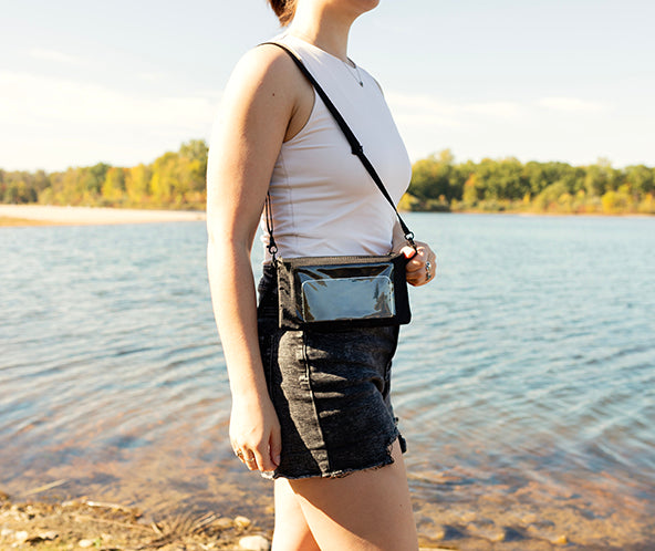Person standing by a lake wearing a white tank top and denim shorts with a clear crossbody bag.